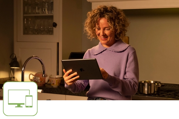 Frau steht in der Küche und schaut auf ein Tablet. Links unten befindet sich ein grünes Icon mit einem Monitor und mit einem Smartphone.