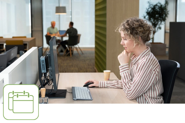 Eine Frau sitzt am Arbeitsplatz und schaut auf ihren Bildschirm. Im Hintergrund sind zwei Männer an einem Besprechungstisch zu sehen. Links unten befindet sich ein grünes Icon mit zwei Kalendern.