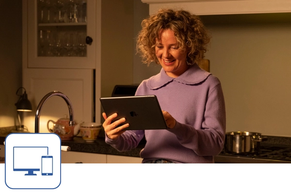 Eine Frau steht in der Küche und schaut lächelnd auf ein Tablet. Links unten ist ein blaues Icon mit einem Monitor und einem Smartphone zu sehen.