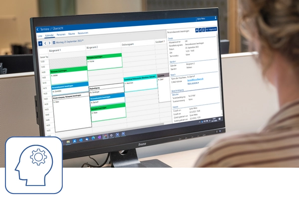 Eine Frau / Sachbearbeiterin schaut auf einen Monitor und hat den Kalender von JCC-Terminvereinbarung geöffnet. Links unten ist ein blaues Icon mit einer Silhouette eines Kopfes und darin ein Zahnrad zu sehen.