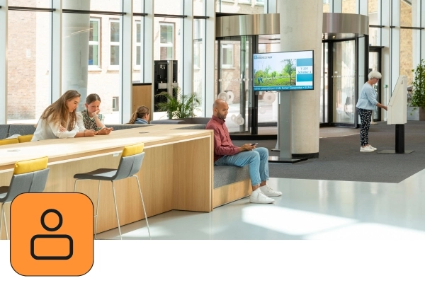 Wartebereich / Eingangsbereich eines Rathauses. Mehrere Bürger warten. Ein Aufrufbildschirm in der Mitte zeigt eine Wartenummer. Rechts holt sich eine ältere Dame am Self-Service-Terminal eine Wartenummer. Links unten ist das orangene Icon von JCC-Besucher zu sehen.